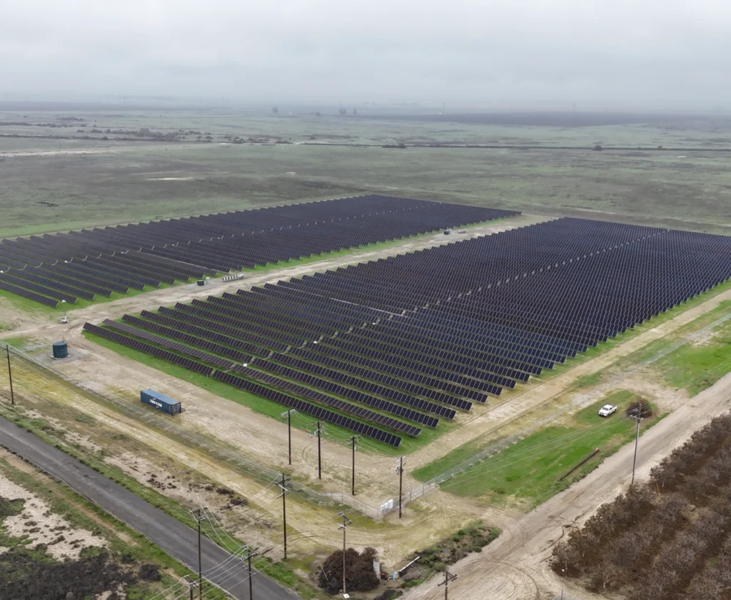 flying-goose-700x574 High-angle aerial view of a large ground-mounted solar panel array situated in a rural field.